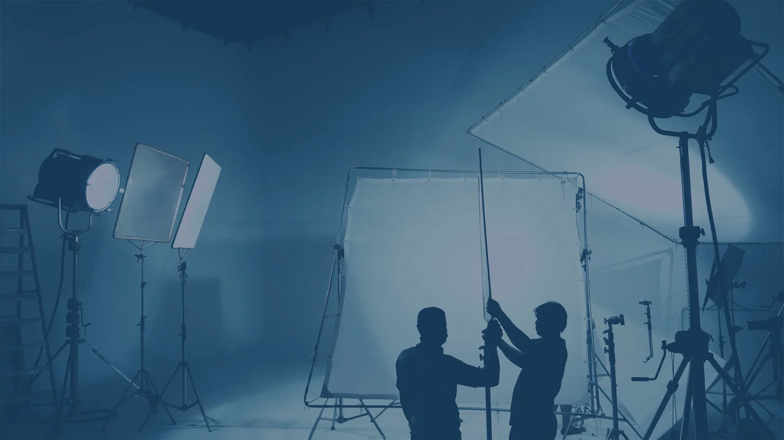 Silhouettes of two people holding a pole in a photography studio with large lights, reflectors, and equipment set against a dimly lit background. . In a demo creation process.